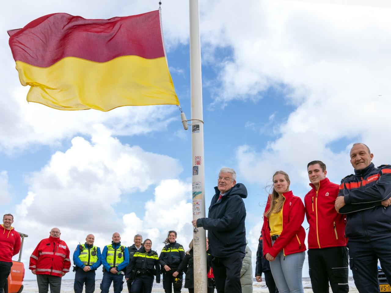 Burgemeester Van Zanen hijst de rood met gele vlag op het strand. Om hem heen staan collega's van de politie, brandweer en lifeguards