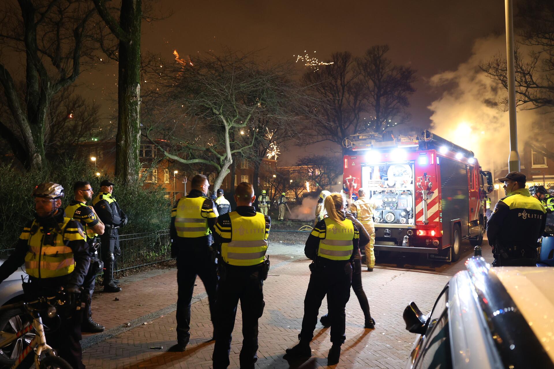 Brandweermannen blussen een brand op oudejaarsavond. Er staat een groep politiemannen omheen. 