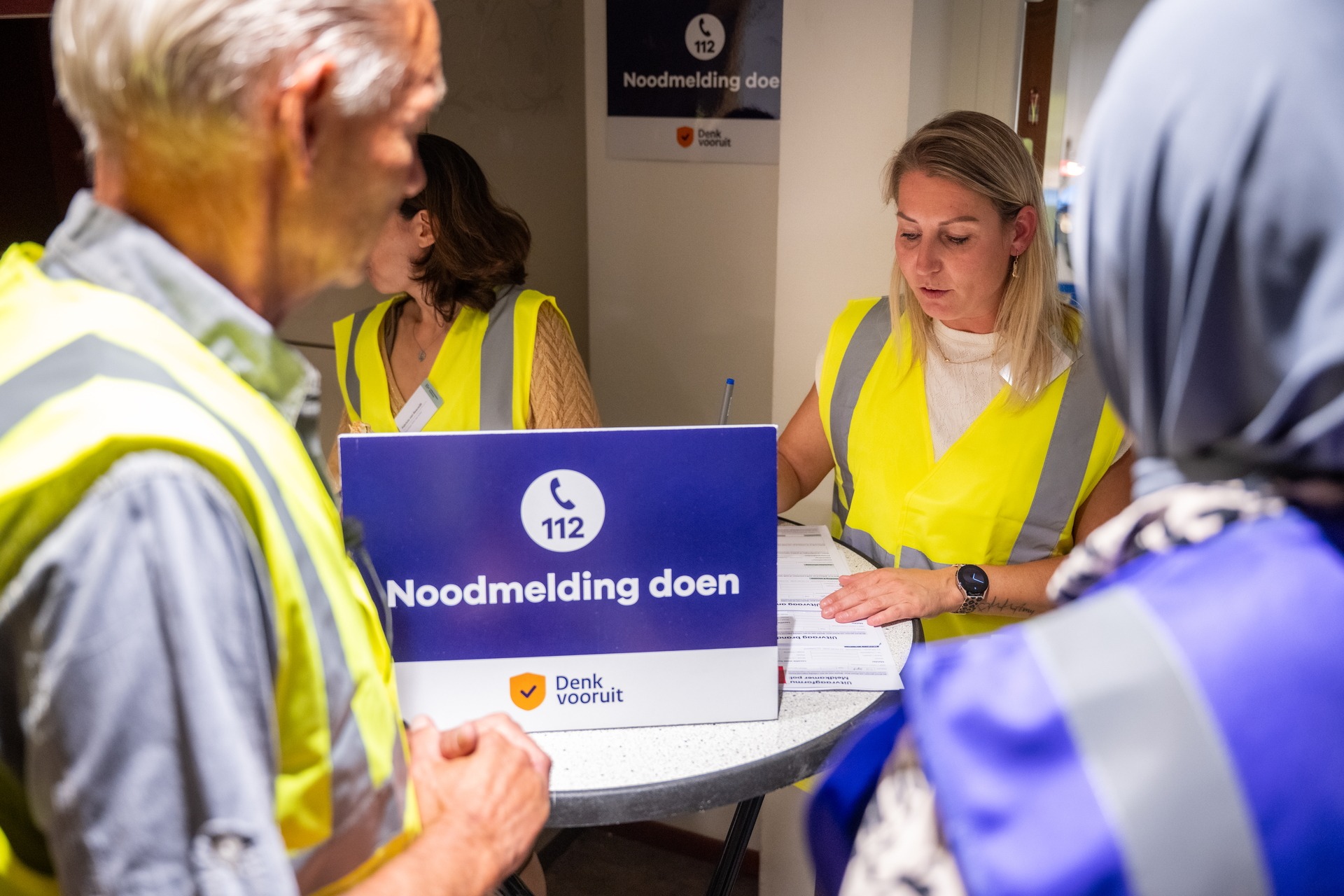 Een foto van mensen bij een balie. Op de balie staat een bord met het woord noodmelding doen