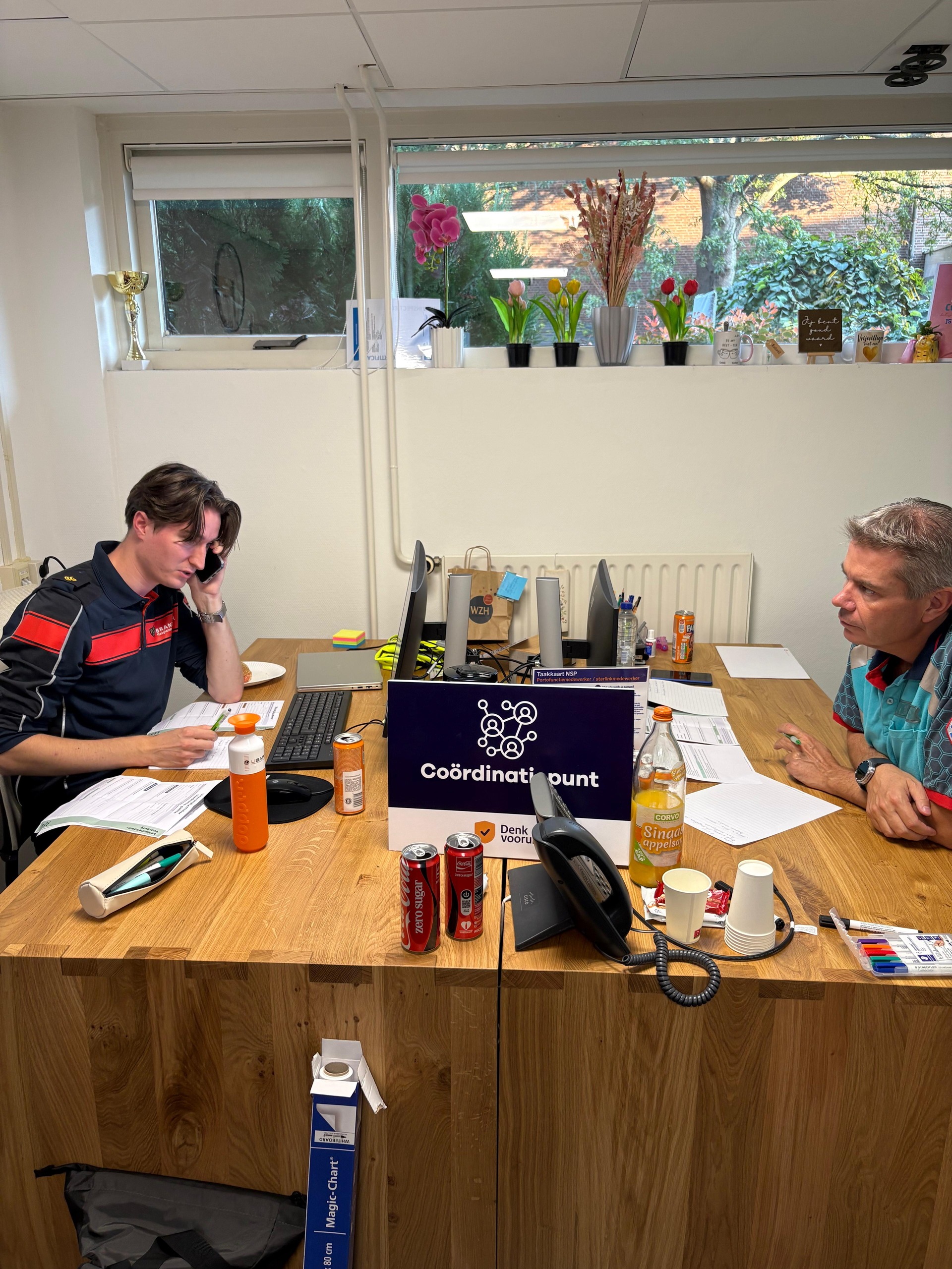 Een foto van twee mannen achter een bureau. Op het bureau staat een bord met de tekst coordinatiepunt