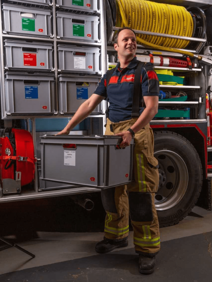 Brandweervrijwilliger Gideon poseert met krat in zijn hand bij een brandweerwagen